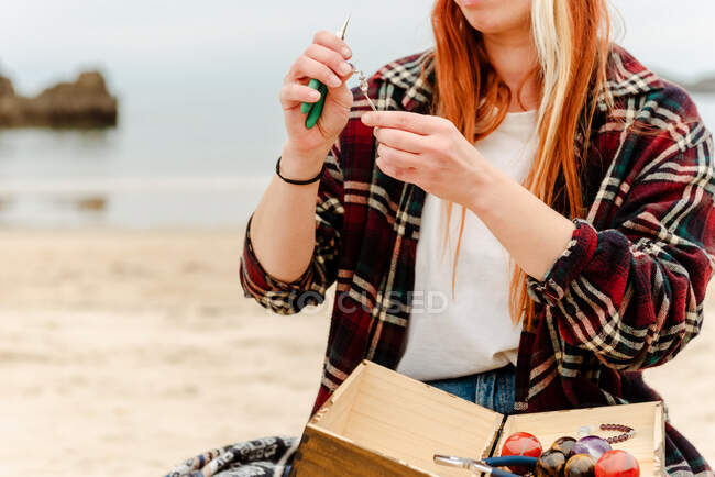Crop anonymous craftswoman creating handmade bijouterie while using semiprecious stones and sitting on beach near sea — Stock Photo