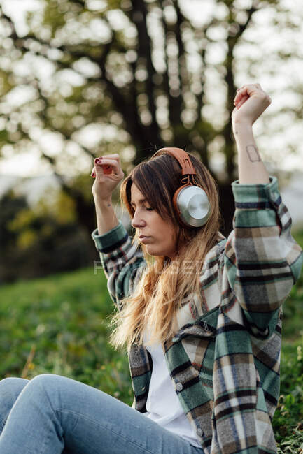 Side view of female listening to music from headphones while having fun sitting with eyes closed on summer meadow against mounts — Photo de stock