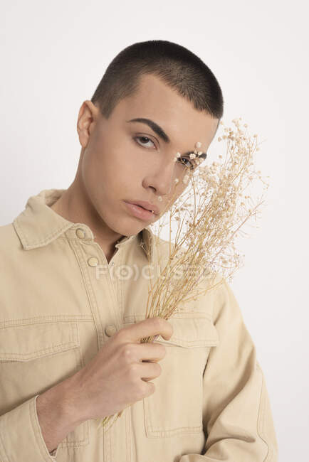 Modello maschio androgeno in camicia alla moda e con mazzo di piante essiccate che guardano la macchina fotografica su sfondo bianco in studio — Foto stock