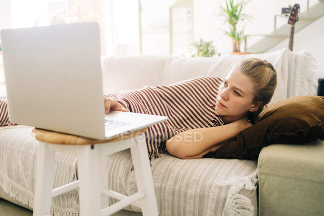 Jovem mulher exausta deitada no sofá e assistindo filme no netbook na sala de estar em casa — Fotografia de Stock