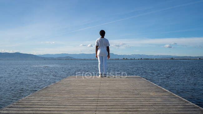 Vue arrière femelle afro-américaine anonyme en vêtements blancs debout sur un quai en bois près d'un vaste lac ondulant sous un ciel bleu clair en été nature — Photo de stock