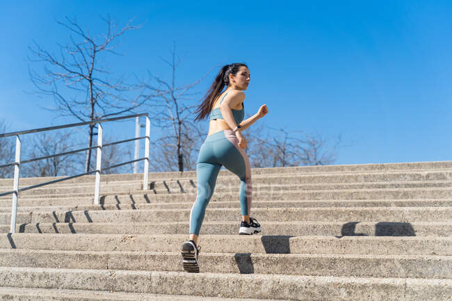 From below back view of fit female runner in sportswear jogging on city staircase and looking away during workout — Stock Photo