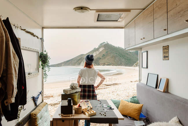 Back view of unrecognizable traveling female artisan standing near truck with handmade accessories on background of sea and mountains — Stock Photo