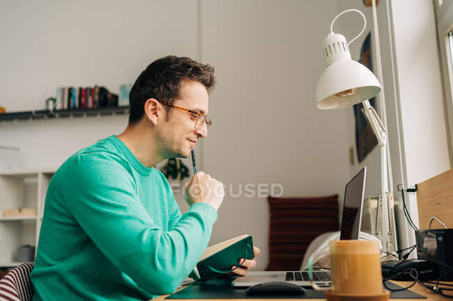 Вид збоку веселий чоловічий віддалений працівник робить нотатки проти нетбука з чорним екраном під час роботи за столом в будинку — стокове фото