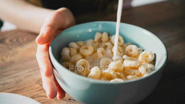 Crop unrecognizable person pouring milk into bowl with delicious corn rings for breakfast on wooden table — Stock Photo