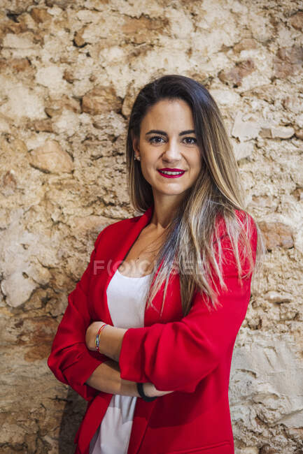 Smiling adult self confident female entrepreneur in red jacket with folded arms looking at camera on brown background — Foto stock