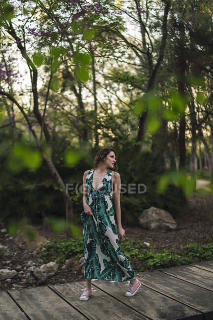Все тело привлекательная молодая женщина в стильном макси-сарае прогуливаясь по набережной в богатом парке и глядя в сторону — стоковое фото