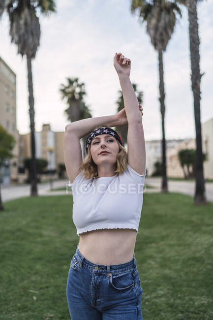 Femme américaine en bandana debout avec les bras levés sur la pelouse avec des palmiers en ville et regardant loin — Photo de stock