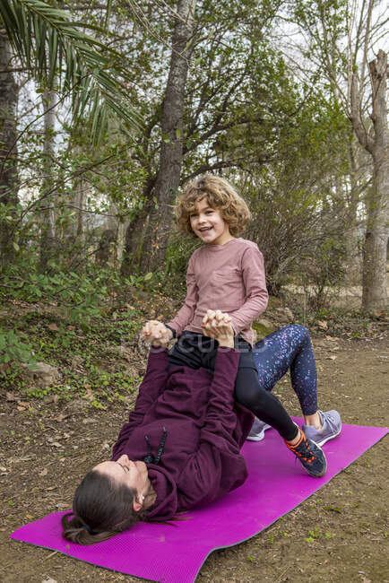 Mother with smiling boy showing Setu Bandha Sarvangasana pose while practicing yoga on mat against trees — Photo de stock