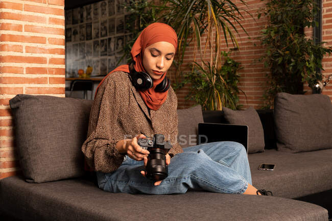 Concentrato giovane donna musulmana in modesto hijab usura e cuffie utilizzando fotocamera professionale foto sul divano accogliente — Foto stock