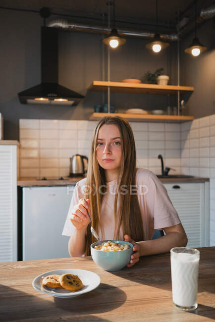 Молода жінка з ложкою і мискою насолоджується смачними кільцями кукурудзи, дивлячись на камеру на кухні — стокове фото