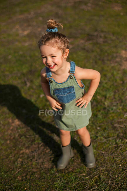 Delighted adorable little girl in overalls standing with hands on waist in meadow and looking away — Stock Photo