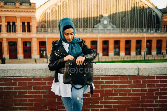 Stylish ethnic female in hijab standing on street and taking things out of leather handbag — Stock Photo