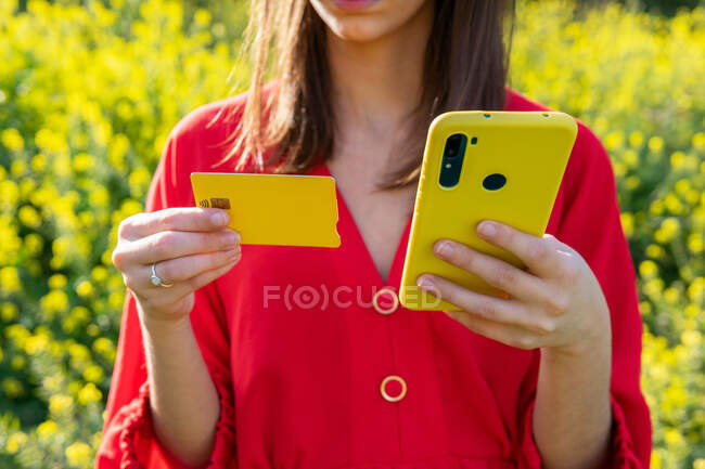 Compradora femenina de cultivos con tarjeta de plástico que paga por la compra en el teléfono celular en el campo en retroiluminación - foto de stock