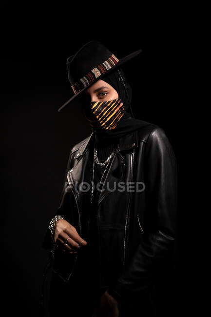 Fashionable Islamic female wearing leather jacket hijab and trendy hat standing looking at camera in dark studio during coronavirus outbreak — Stock Photo