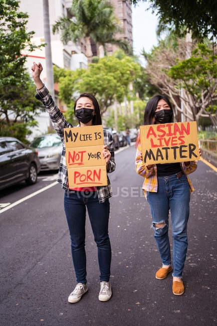 Ethnic females in masks holding posters protesting against racism in city street and looking away — Stock Photo