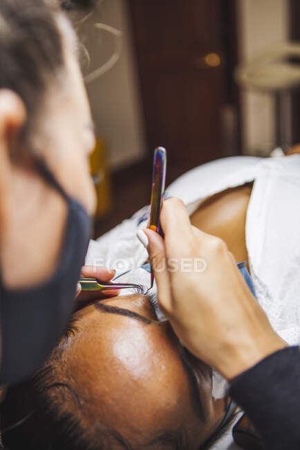 Crop unrecognizable cosmetologist with tweezers applying fake eyelashes for extension on eye of ethnic client with face protective mask in salon during coronavirus pandemic — Fotografia de Stock
