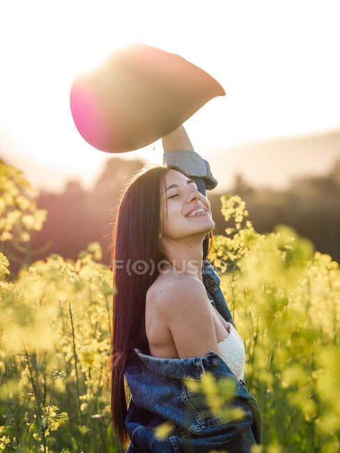 Side view of delighted young brunette with eyes closed in white top hat and lowered jacket holding hat and laughing merrily on blooming rapeseed field on sunny day — Stock Photo