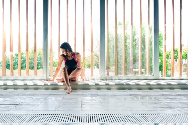 Молодая одинокая спортсменка в купальниках, сидящая со скрещенными ногами на полу после тренировки под солнцем — стоковое фото