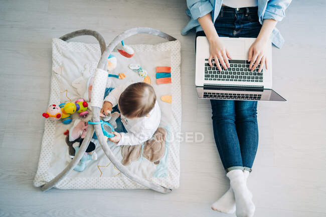Vista superior madre anónima con traje casual sentado en el suelo con netbook en vueltas y extendiendo la mano a lindo bebé jugando con juguetes en el suelo - foto de stock