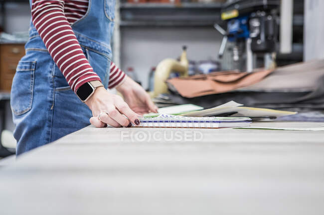 Vista lateral de bajo ángulo de cultivo mujer artesana anónima de pie con bloc de notas y telas surtidos en el banco de trabajo en el taller de tapicería de motocicletas - foto de stock