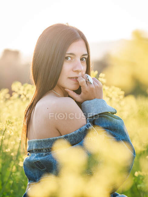 Jovem morena em top branco e jaqueta de ganga abaixada olhando para a câmera no campo de colza florescendo no dia ensolarado — Fotografia de Stock