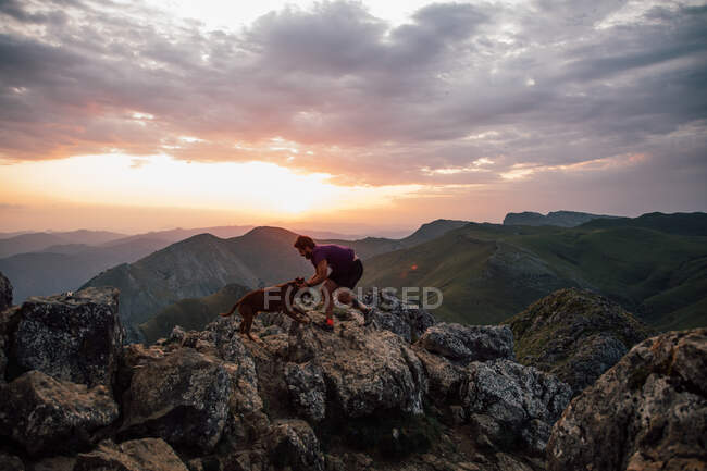 Hombre acariciando perro leal mientras está de pie en la roca de la cresta de la montaña bajo el cielo nublado puesta del sol - foto de stock