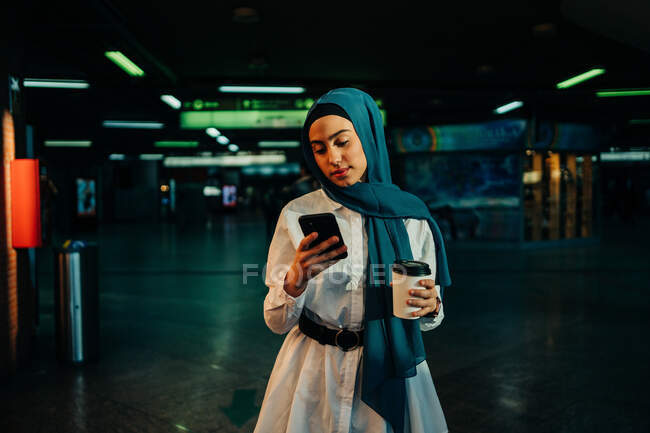 Elegante donna musulmana in hijab in piedi con caffè da asporto in metropolitana e navigare in Internet sul cellulare — Foto stock