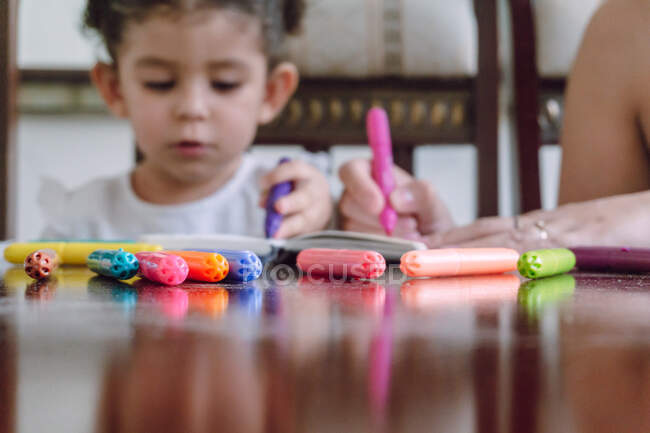 Cultivo mujer irreconocible y niño pequeño sentado a la mesa y dibujo con marcadores en el cuaderno - foto de stock