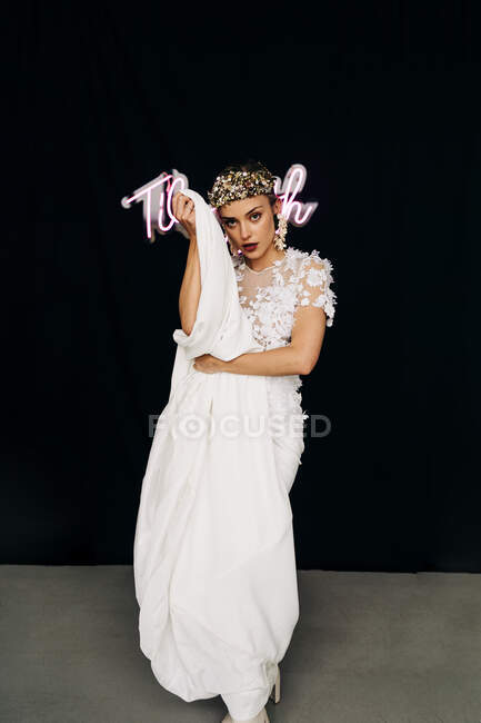 Charming young female model in bohemian elegant white lace gown and floral wreath standing against black background with neon inscription — Stock Photo