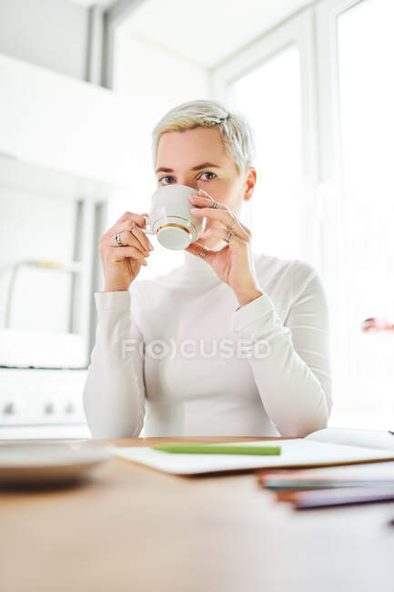 Жінка-астролог п'є гарячий напій з чашки, дивлячись на камеру вдома на сонячному світлі — стокове фото