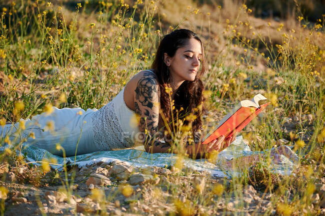 Dreamy charming brunette in white dress lying on field meadow and reading book in sunlight — Foto stock