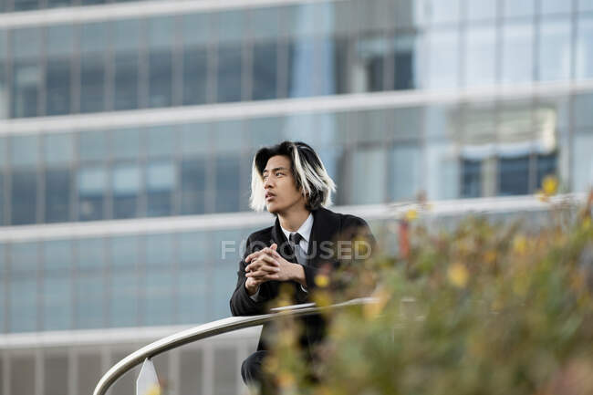 Young ethnic male entrepreneur in coat leaning on fence while looking away against contemporary city house — Stock Photo
