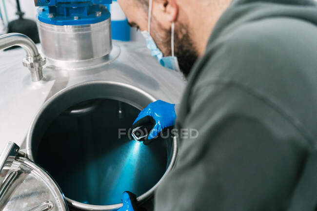 Von oben sieht man einen männlichen Ingenieur mit Blaulicht bei der Überprüfung eines Metallgefäßes in einer Brauerei während einer Pandemie — Stockfoto