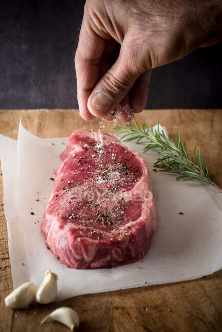 Crop anonymous male cook seasoning uncooked meat piece with salt and ground black pepper between garlic cloves and fresh rosemary sprig — Stock Photo