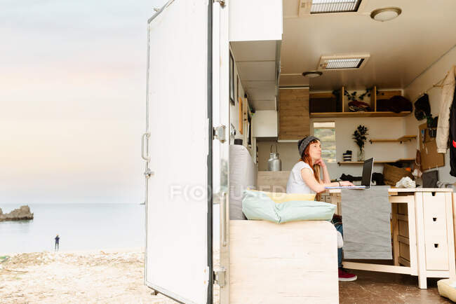 Vista lateral del viajero femenino enfocado sentado en la mesa en camión y tomando notas mientras trabaja en el proyecto de forma remota y apoyándose en la mano - foto de stock