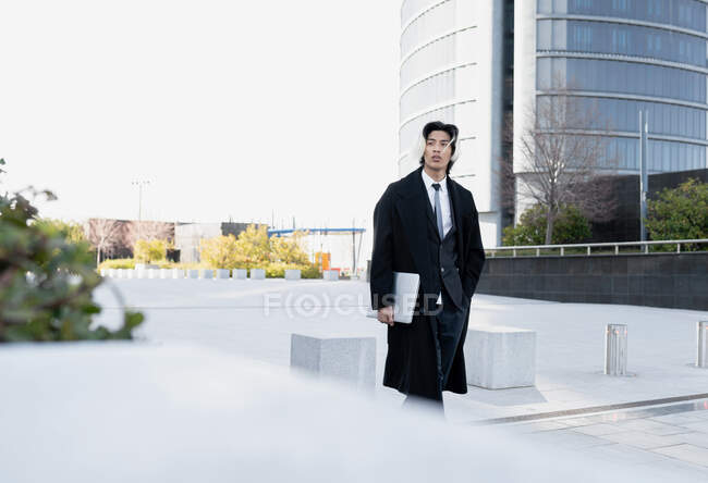 Young ethnic male executive in coat with hand in pocket and netbook looking away while strolling in city — Stock Photo