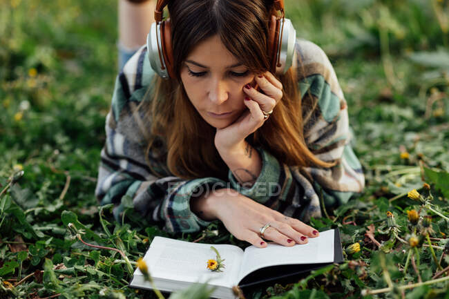 Young attentive female in modern headset reading textbook while lying on meadow in summer — Photo de stock
