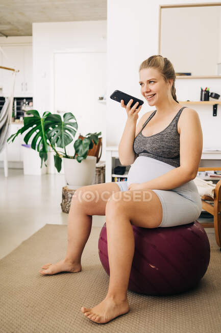Young content expectant female sitting on fitness ball while sending voice message on cellphone and looking away at home — Stock Photo