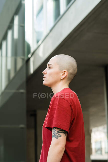 Vue latérale de la jeune lesbienne non émotionnelle en t-shirt rouge regardant la caméra contre la façade de la maison en ville — Photo de stock