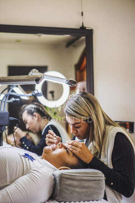 Female beauty master with tweezers applying fake eyelashes on face of ethnic client in salon — Photo de stock