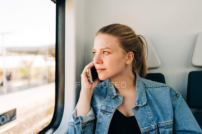 Young casual female in denim jacket having phone call looking away in window of train while commuting — Stock Photo