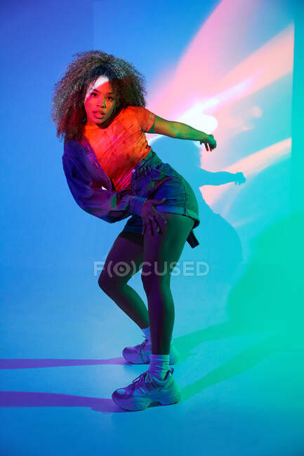 Cuerpo completo atractivo ajuste afroamericano femenino en pantalones cortos bailando en luces de neón en el estudio y mirando a la cámara - foto de stock