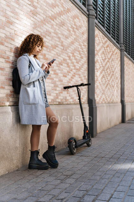 Side view of young African American female with curly hair wearing blue coat browsing on mobile phone while standing against brick wall and looking at screen with smile — Foto stock