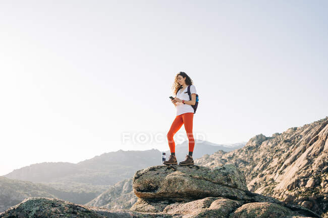 Ganzkörper positiver junger Reisender mit lockigem, dunklem Haar in lässiger Kleidung, der beim Wandern in den Bergen am sonnigen Tag mit dem Handy lächelt — Stockfoto