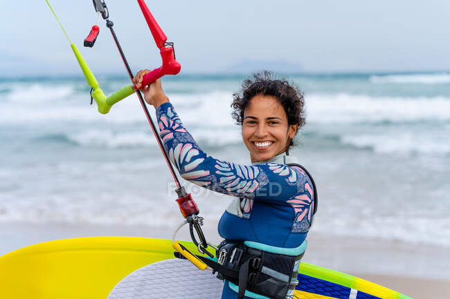 Alegre kiter femenino en traje de neopreno sosteniendo la barra de control mientras mira la cámara en la orilla del océano arenoso - foto de stock