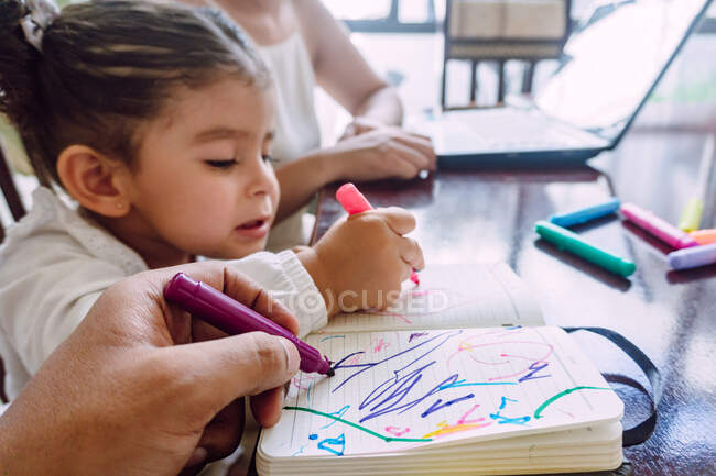 Crop unrecognizable woman browsing laptop while little child sitting at table and drawing with markers in notebook — Stock Photo