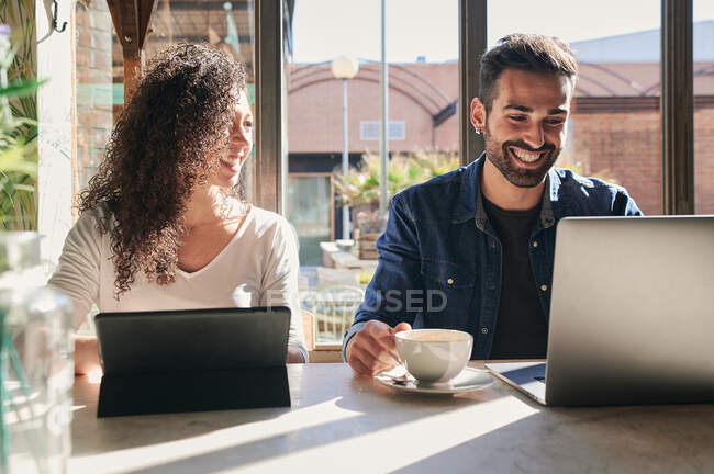 Funcionário remoto masculino étnico desfrutando de bebida quente contra laptop e parceiro feminino com tablet à mesa à luz do sol — Fotografia de Stock
