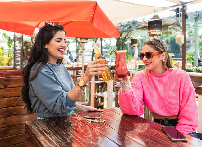 Felice giovani amiche in abiti casual clinking bottiglia di birra e bicchiere di cocktail di bacche mentre seduto a tavola in soleggiato bar all'aperto — Foto stock