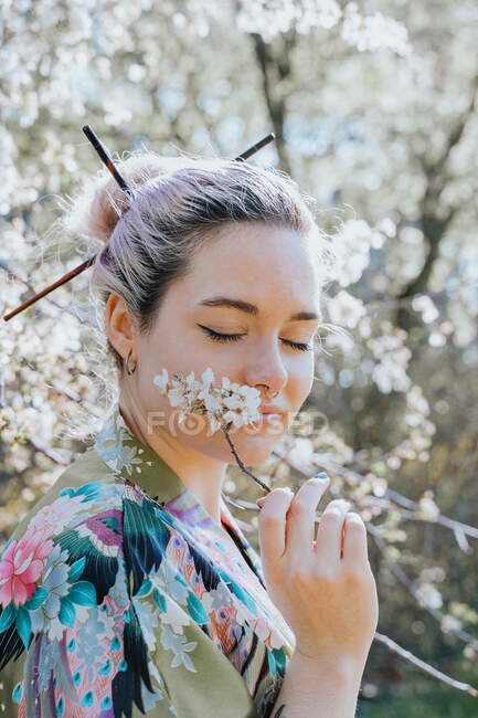 Side view of young female in traditional kimono holding blossoming flower sprig in garden on sunny day looking at camera — Stock Photo